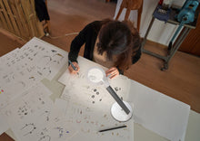 Load image into Gallery viewer, Person working on a design project with sketches and a Halo Go Magnifying lamp in a workshop setting