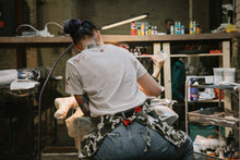 Load image into Gallery viewer, Woman's back as she tattoo's a client using the Electra lamp to provide light