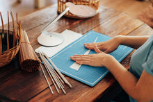 Load image into Gallery viewer, Person cutting paper on a blue cutting mat with tools on a wooden table, using the light from a Halo Go lamp.
