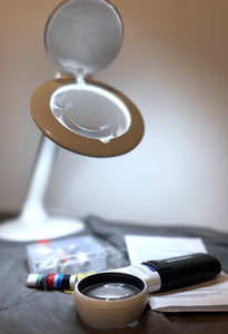 Halo magnifying lamp and Handheld round magnifier resting on a piece of fabric with sewing supplies. 