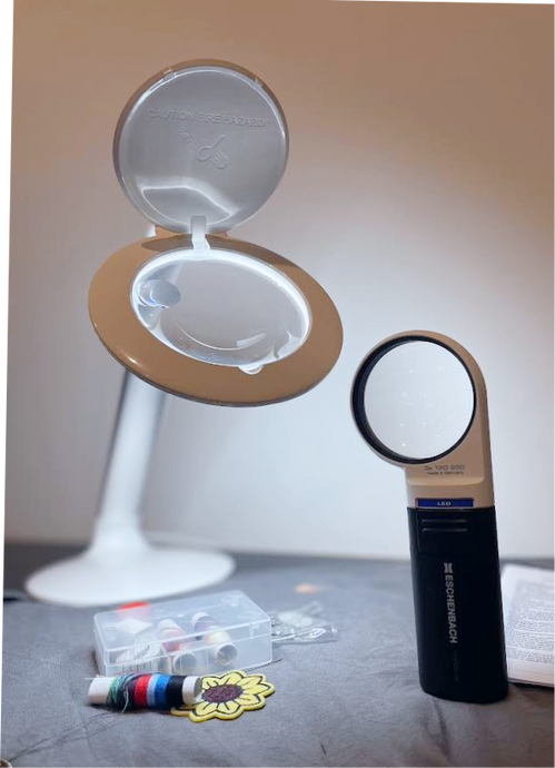Halo magnifying lamp and Handheld round magnifier resting on a piece of fabric with sewing supplies. 
