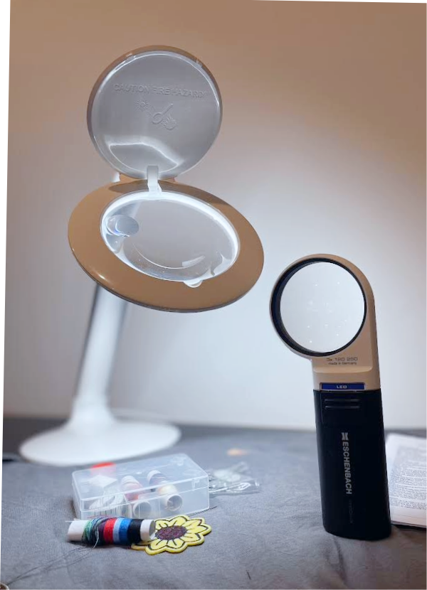 Halo magnifying lamp and Handheld round magnifier resting on a piece of fabric with sewing supplies. 
