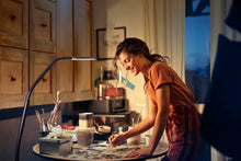 Load image into Gallery viewer, Woman painting at a round table, using the Electra lamp to light her work space.
