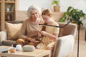 Woman and young girl sitting on a sofa, the woman is teaching the child to knit using the Electra lamp for light